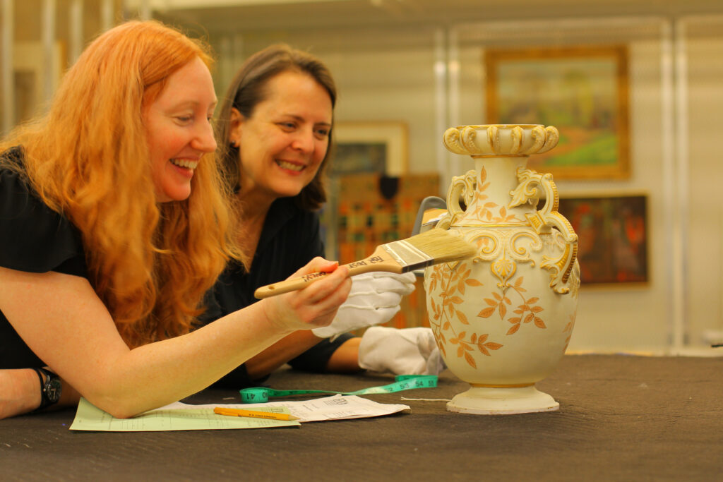 two women dust a vase with paintbrushes.