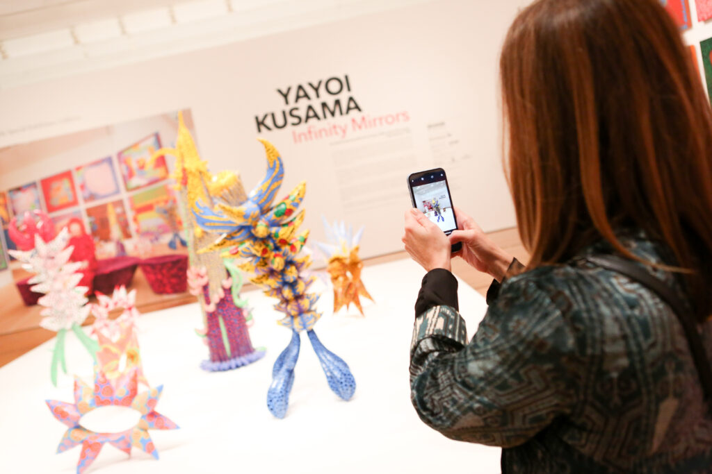 A woman with brown hair holds an iPhone to take a photo of colorful sculptures in an art exhibition