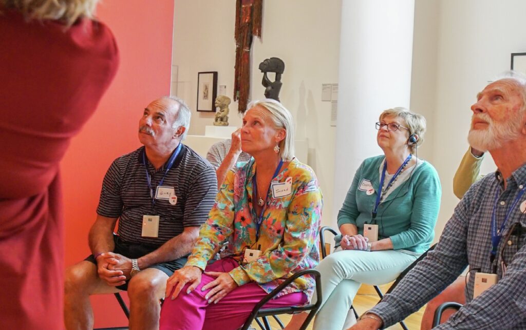Musing Together A group of people sit in chairs, attentively listening to a speaker in a brightly lit room with artwork on the walls.