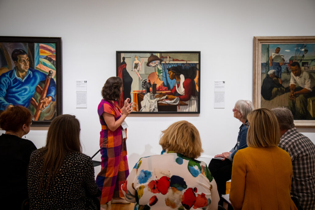A group of people sitting on stools in an art gallery, listening to a woman in a colorful dress explaining a painting. The gallery features several large paintings displayed on white walls.
