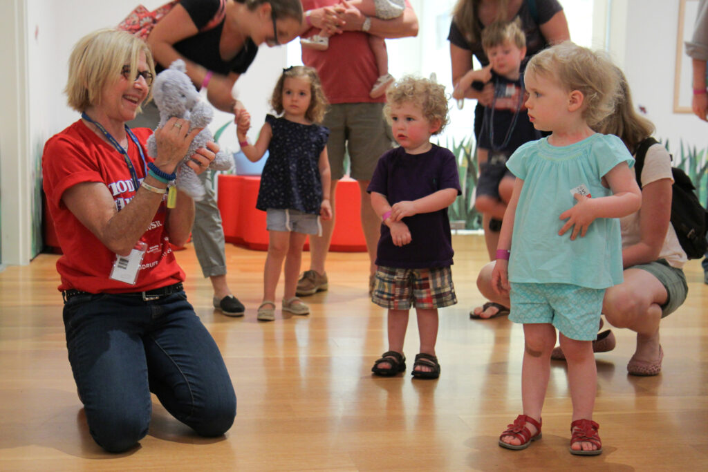 Toddlers stand mesmerized by a woman holding up a stuffed animal in the High Museum galleries.