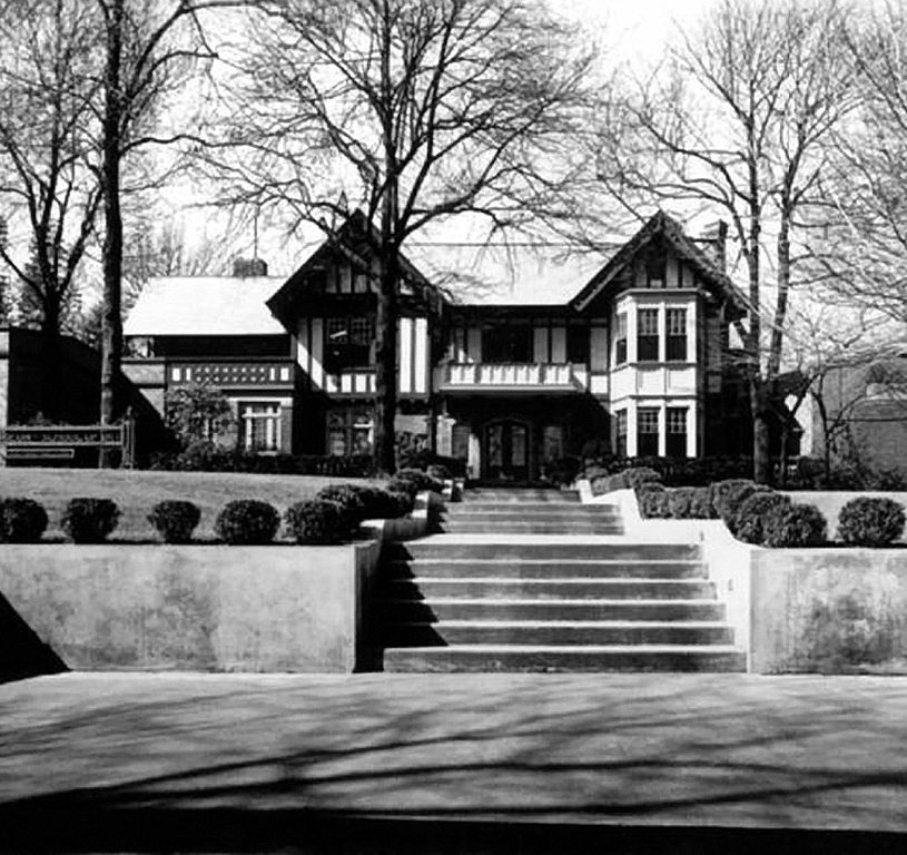 Wide concrete steps lead from the sidewalk to a two story Tudor home.