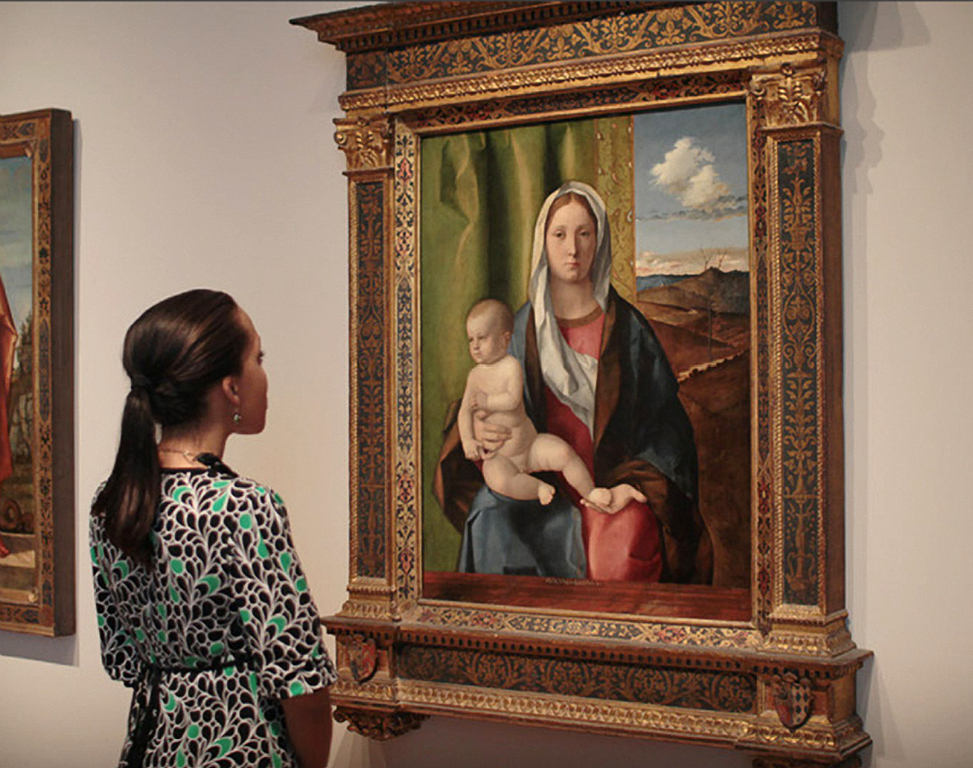 A woman with long brown hair pulled back in a ponytail gazes at a sixteenth century portrait of the Madonna and child by Giovanni Bellini.
