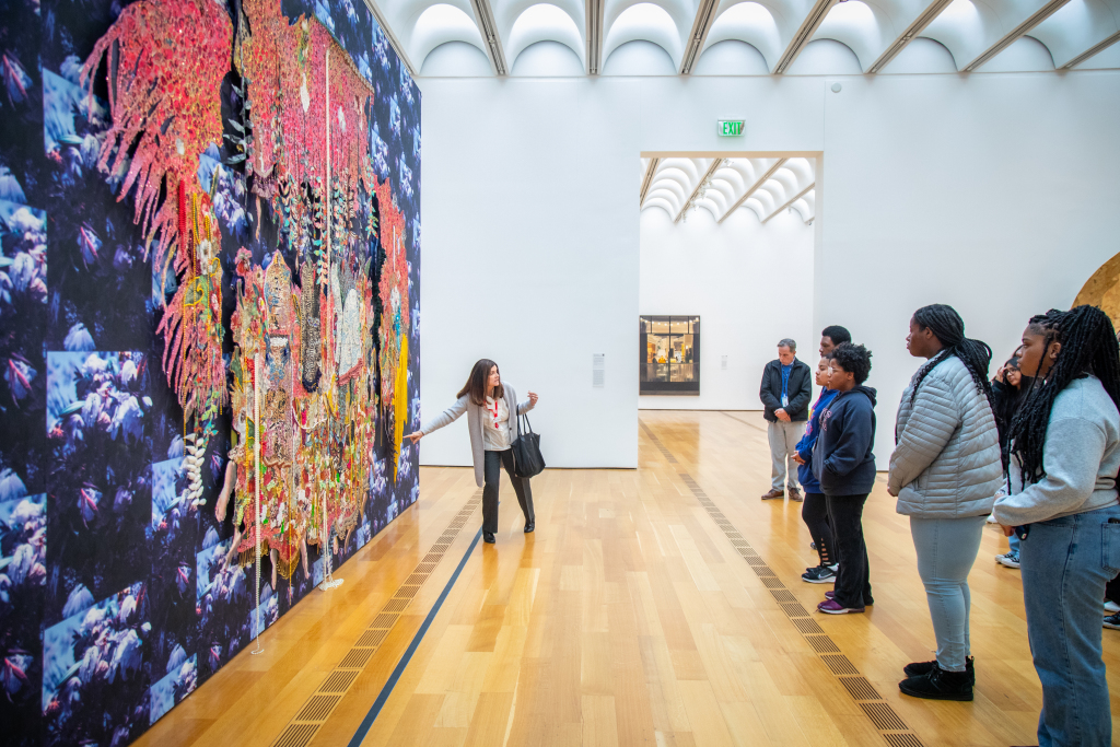 Docent points to a large collage of wallpaper, colorful beads, and other mixed media as a group of students listen.