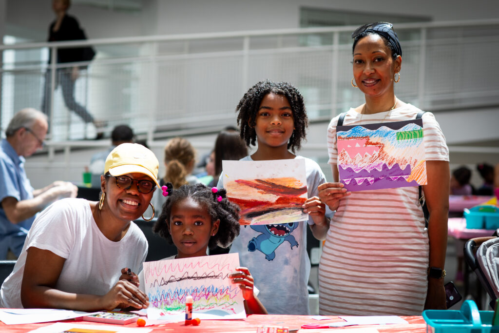 Two woman and two young children smile while holding up drawing artworks