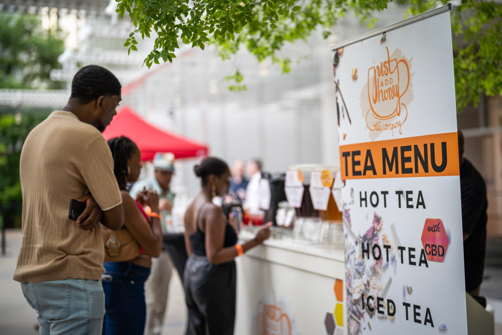 Just add honey, High frequency Friday People stand in line at an outdoor tea stand with a banner displaying the tea menu, including hot tea, hot tea with CBD, and iced tea. Trees and a red tent are visible in the background. One man pays at the counter as others wait to place their orders.