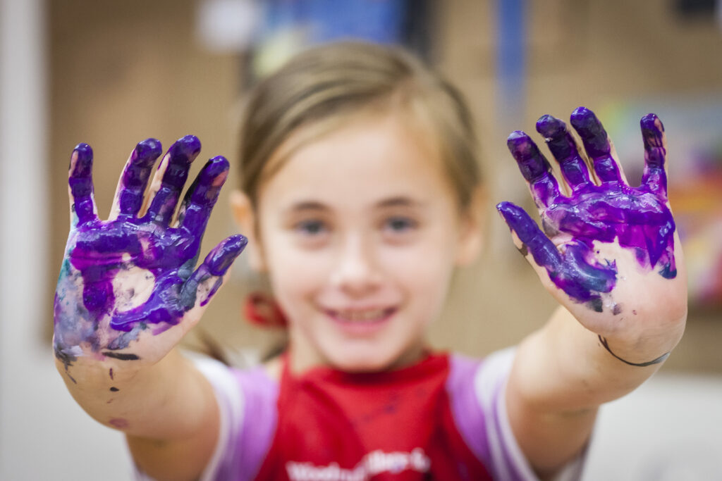 young girl with blonde hair holds up two hands covered in purple paint.