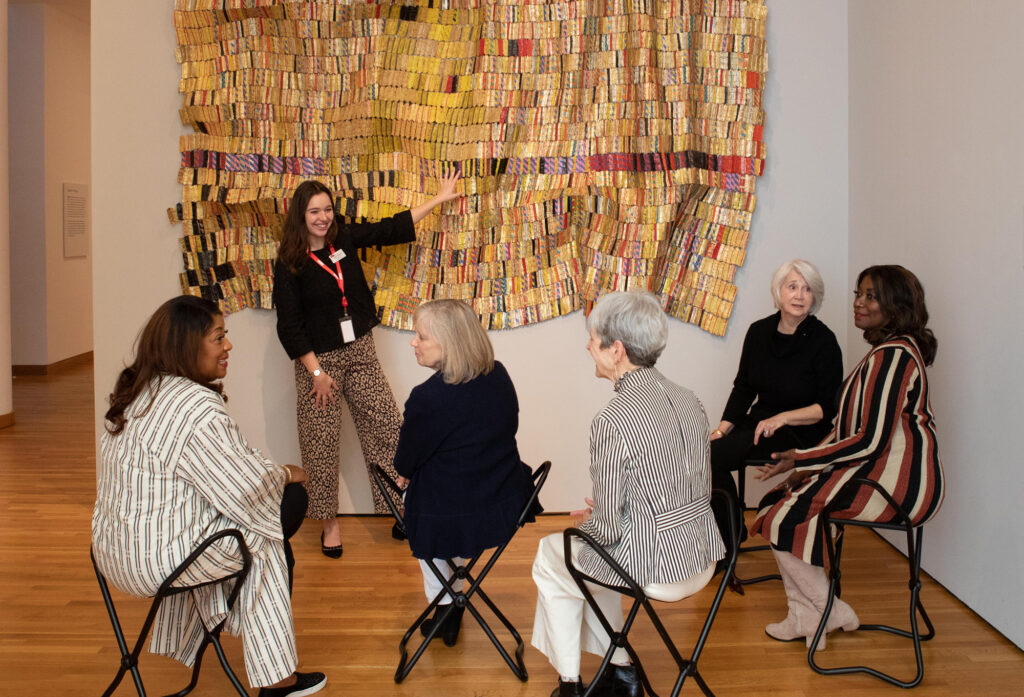a group of older women sit on folding chairs in front of a shimmering golden tapestry of metal bottle wrappers as a young woman points out details in the artwork.