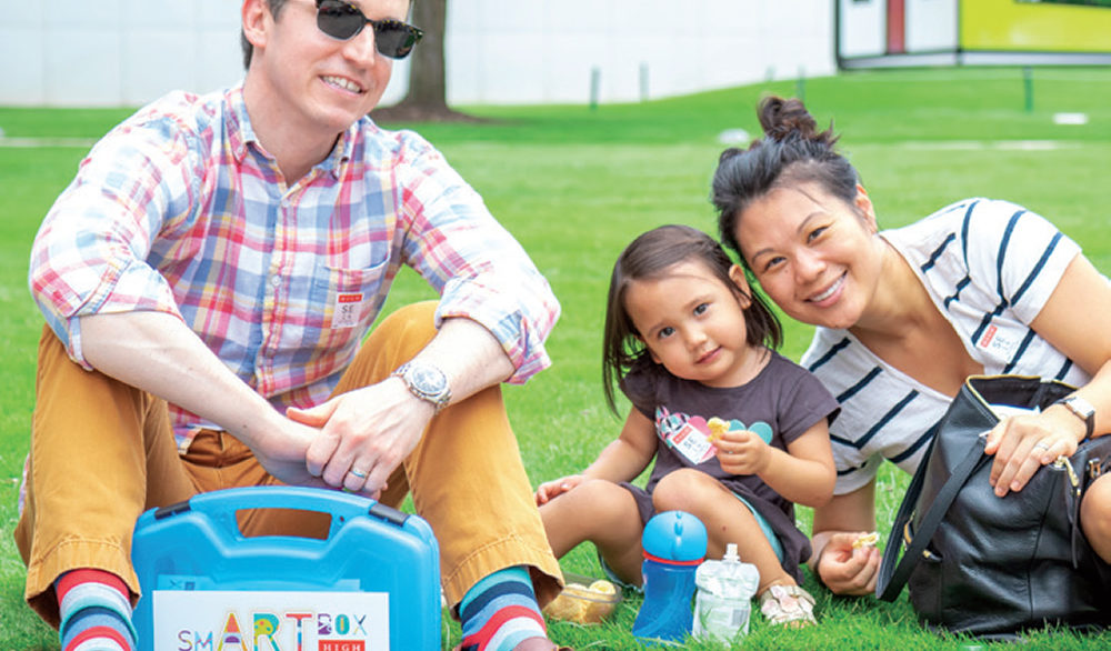 A man, woman and their toddler daughter sit on a bright green lawn in front of the High Museum.